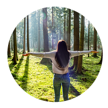 Beate Kammerbauer mit ausgebreiteten Armen von hinten zu sehen im Wald, umgeben von Sonnenstrahlen und Bäumen.