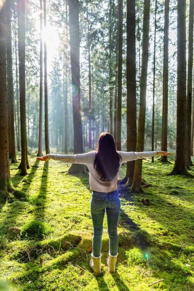 Frau mit ausgebreiteten Armen in einem sonnendurchfluteten Wald.