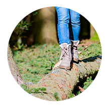 Person in Jeans und Wanderstiefeln balanciert auf einem Baumstamm im Wald.