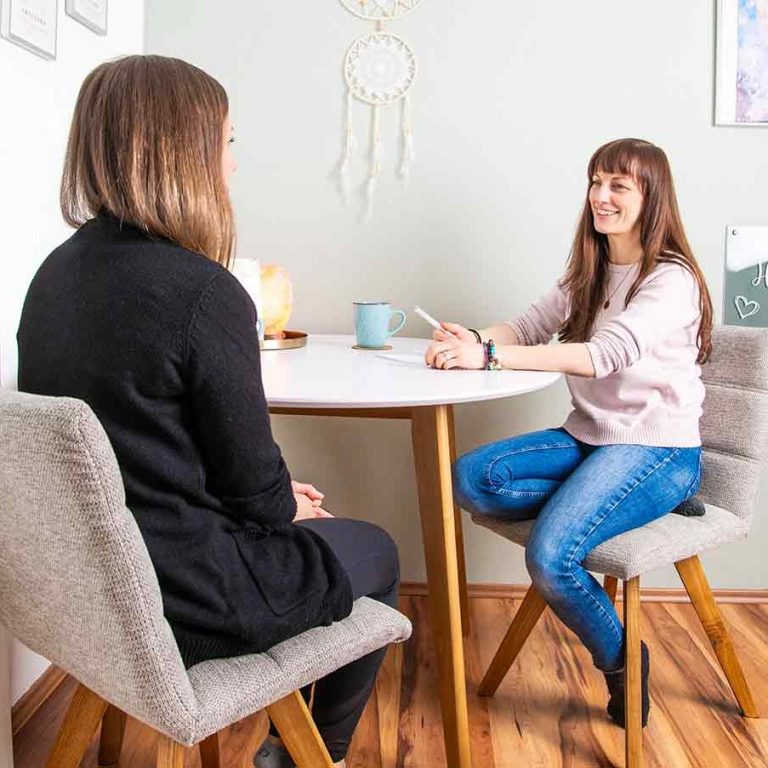 Beate Kammerbauer sitzt mit einer Klientin am Tisch, lächelt und hat einen Stift in der Hand. Die beiden kommunizieren.