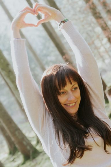 Frau mit langen Haaren bildet mit den Händen ein Herzzeichen in einem Wald.