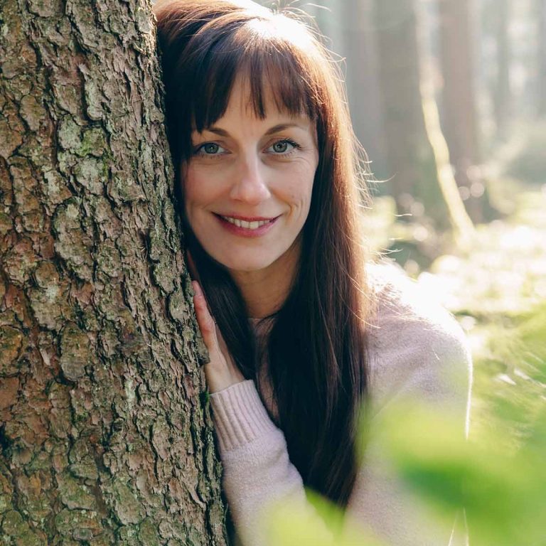 Beate Kammerbauer (Frau mit langen, braunen Haaren) die an einem Baum in einem Wald lehnt und lächelt.